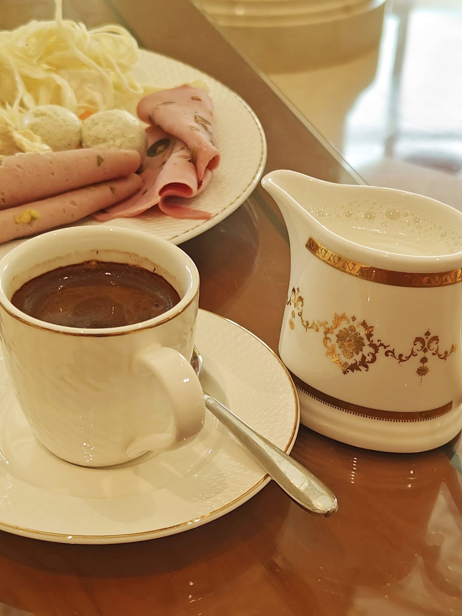 Cup of traditional Turkish coffee served in an ornate gold-trimmed cup and saucer on a breakfast table at Raffles The Palm Dubai, with pastries and fruit softly blurred in the background.