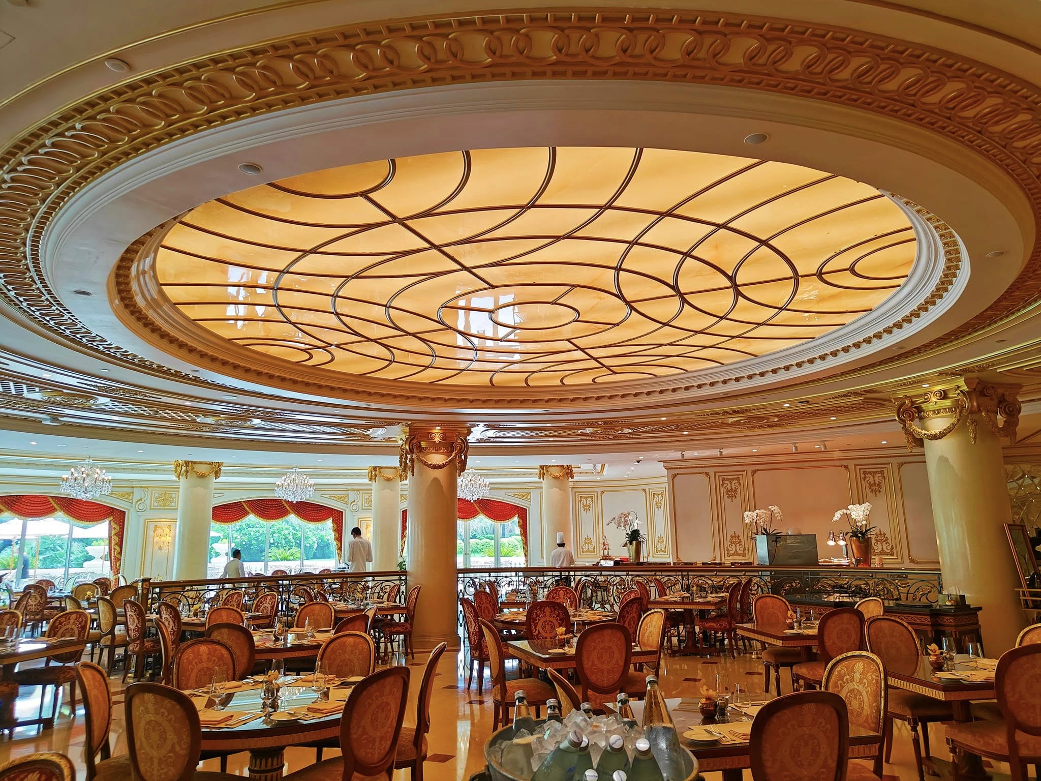 Elegant restaurant interior at Raffles The Palm Dubai featuring crystal chandeliers, plush seating, gold accents, and large windows creating a refined, European-inspired dining atmosphere.