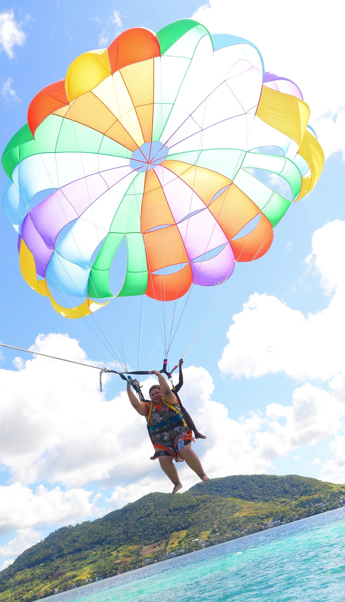 A traveller parasailing high above the turquoise lagoon off the south coast of Mauritius, with the island's lush coastline stretching below